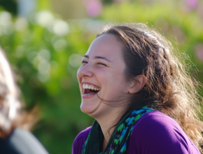 Woman laughing joyfully outdoors in warm sunlight with eyes crinkled in genuine delight, demonstrating the authentic expression and therapeutic benefits of laughter yoga, with natural greenery in the background creating a serene practice environment