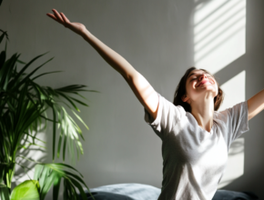 Woman stretching her arms in morning sunlight by a window, with eyes closed and a peaceful smile, demonstrating the gentle backbend stretch in a bright room with a houseplant, embodying the calm and rejuvenating benefits of a morning stretch routine